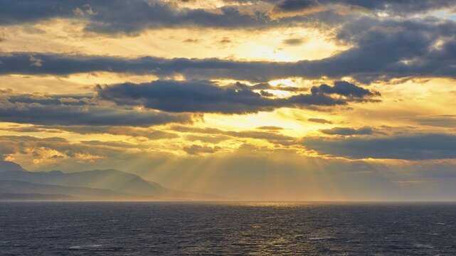 The sun sends its rays through the clouds over the sea near a mountain, Cape Melagkavi, Gulf of Corinth, Corinth, Peloponnese, Greece