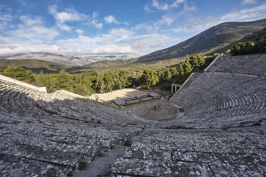 Panoramic view of an ancient amphitheatre against a mountain backdrop under a partly cloudy sky, Ancient Amphitheatre, Epidaurus, Peloponnese, Greece