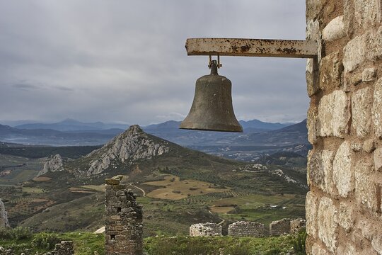An ancient bell hangs against a backdrop of ruins and a mountainous landscape with cloudy skies, Archaic Castle, Akrokorinthos, Corinth, Peloponnese, Greece