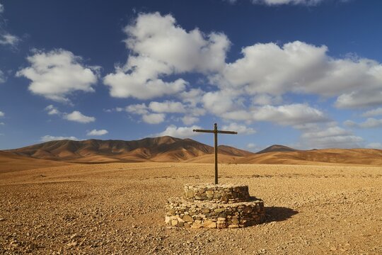 Island interior, barren landscape, wooden cross on stone pedestal, red-brown barren hills, blue sky, white clouds, Fuerteventura, Canary Islands, Spain