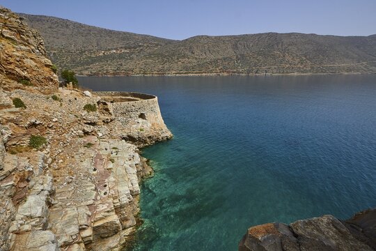 Rustic stone wall on cliffs above a turquoise sea with mountains in the distance, Venetian sea fortress, Leprosy Island, Spinalonga, Elounda Bay, Mirabello Bay, East Crete, Crete, Greek Islands, Greece