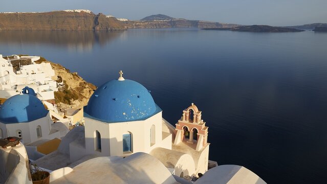 View down to white church with blue dome, view from Oia over the caldera to the cliff, Imerovigli, Firostefani, Fira, villages on the cliff, sea in the caldera smooth as glass and dark blue, sky light blue and cloudless, Oia, Santorini Island, Cyclades, Greece