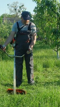 Man in gray overalls operates a string trimmer to cut grass in a lush garden surrounded by trees and greenery on a sunny day