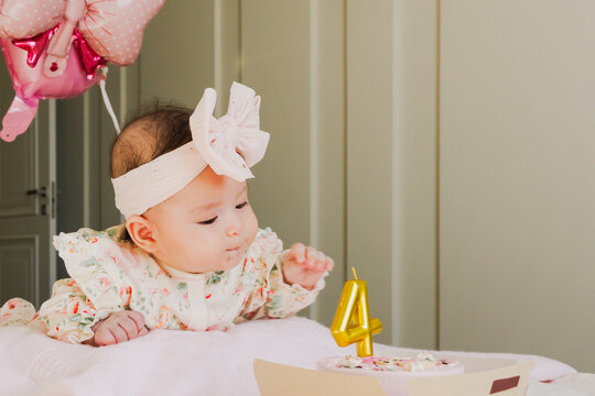 Baby girl in floral dress with large bow reaches for birthday cake with number four candle, pink balloon in the background, soft indoor setting with neutral colors