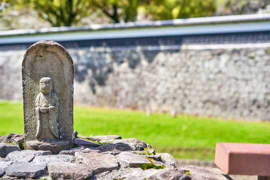 A close-up view of an ancient weathered stone Jizo statue, a Buddhist guardian deity, carved in relief within an arched stele and positioned on a moss-covered stone wall. The statue shows signs of