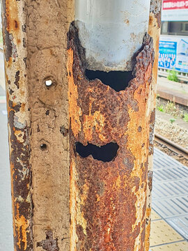 Close-up of a severely rusted metal utility pole at a train station in Japan, showing extensive corrosion damage. The weathered surface features peeling beige paint revealing orange-brown rust