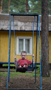 Candid moment of young kid on blue swing in pine forest park. Casual summer day, barefoot boy gazing at viewer. Ideal for nature and childhood projects.