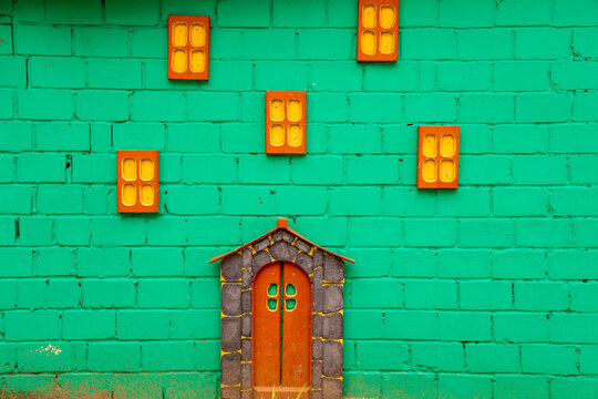 A vibrant green brick wall features a decorative installation mimicking a fairy tale house facade with miniature orange windows and a small stone-trimmed entrance door.