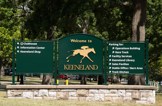 A green 'Welcome to Keeneland' entrance sign in Lexington, Kentucky, featuring a running horse logo and parking directions at the historic Thoroughbred race track.