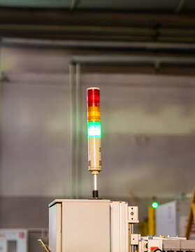 A glowing industrial signal tower light with red, yellow, and green indicators stands mounted atop a machinery control box within a manufacturing facility.