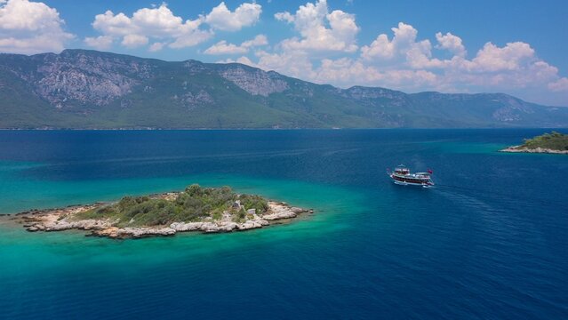 A small island off the coast of Sedir Adası. A tour boat heading towards Akyaka,  lush green mountains rise under a white cloudy sky on the background. Aerial view. Gulf of G&ouml;kova, Muğla, Turkey