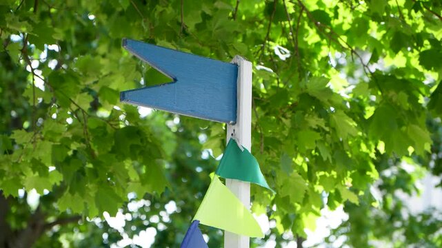 A simple blue wooden swallowtail flag acting as a weather vane or directional sign stands on a white post decorated with colorful small triangular bunting banners
