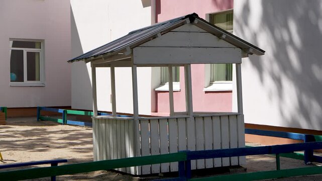 A small, white wooden gazebo structure with a gabled roof stands inside an empty playground sandbox area next to a building on a sunny day.