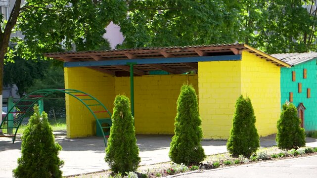 A bright yellow brick playground pavilion with a brown roof stands behind a neat row of small green thuja trees on a sunny summer day.