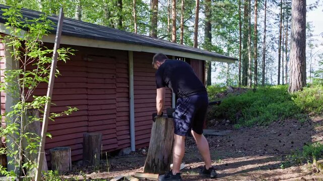 A man splits logs with an axe on a wooden chopping block outside a red wooden shed in a forest. 60Fps.