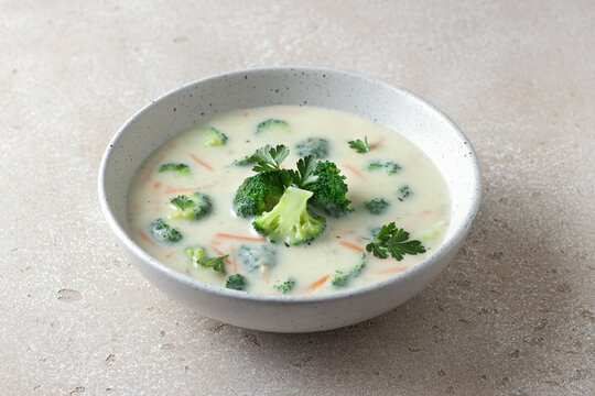 Homemade creamy broccoli soup with potatoes carrots and parsley in ceramic bowl on light textured background