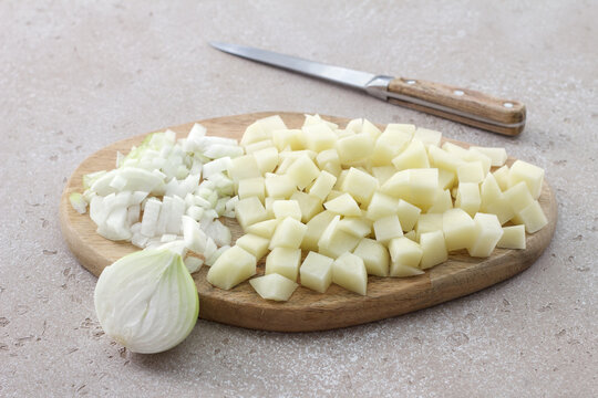 Fresh diced potatoes and chopped onion on a wooden cutting board with a kitchen knife, ready for homemade soup cooking on a light textured background