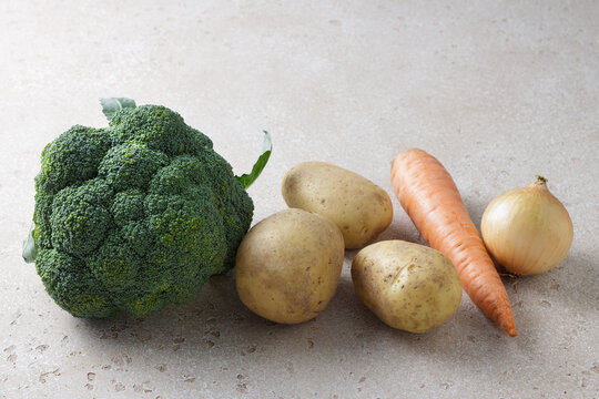 Fresh raw broccoli, potatoes, carrot and onion on light stone background. Healthy vegetables and cooking ingredients for homemade meals and soups