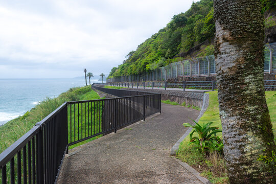 MIYAZAKI, JAPAN: A concrete pedestrian path with a black metal handrail running parallel to the tiered coastal highway and its protective rockfall netting at Horikiri Pass (Horikiri-toge) in Kyushu.