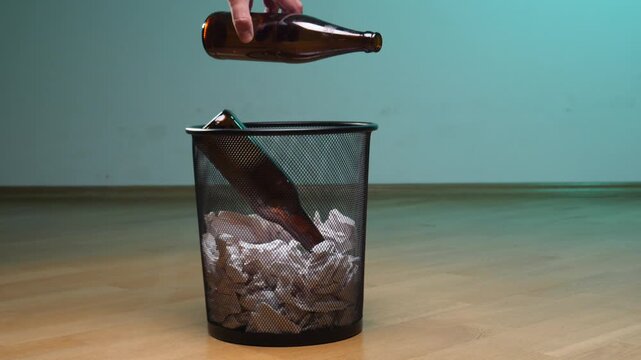 Hand dropping a brown glass bottle into a black mesh trash can filled with crumpled paper, symbolizing recycling glass, responsible waste disposal, and environmental conscience