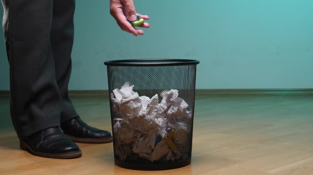 Hand improperly disposing of spent aa batteries into an office rubbish bin filled with crumpled paper, highlighting environmental disregard for proper recycling and hazardous waste management