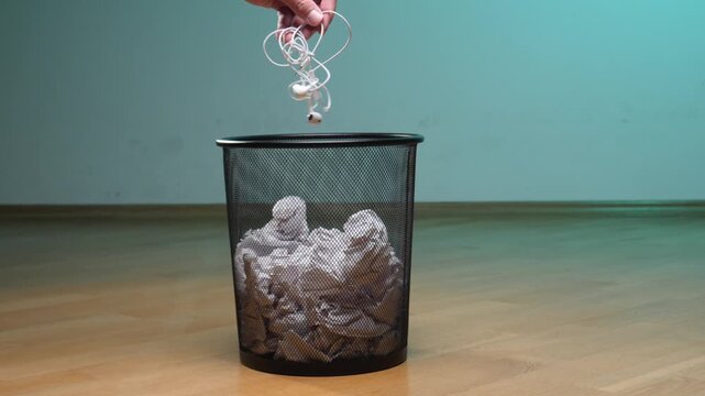Hand holding tangled white wired earphones, placing them over a black mesh trash bin filled with crumpled paper, symbolizing electronic waste disposal and technological obsolescence