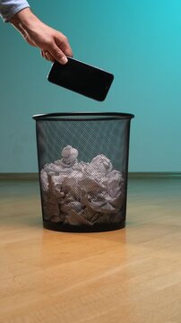 Vertical video human hand dropping an old smartphone into a black mesh trash can filled with crumpled paper. Symbolizing digital detox. Electronic waste
