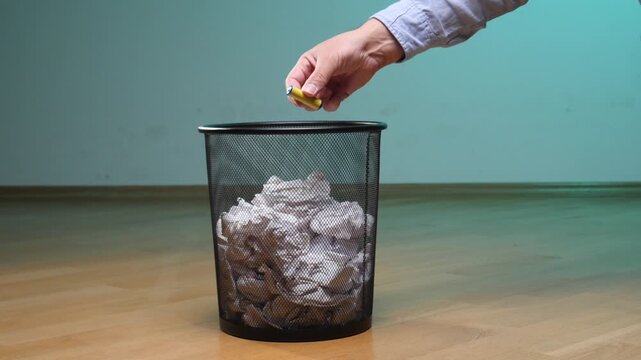 Hand dropping a used alkaline battery into a waste bin filled with crumpled paper, illustrating the incorrect disposal of hazardous waste and promoting environmental awareness