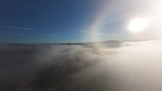 Cinematic aerial drone footage flying through dense morning fog over a peaceful mountain village with small lakes and lush forest. Low clouds cover the valley while the drone rises above the mist