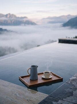 Floating wooden tray with steaming coffee and ceramic pitcher in an infinity pool overlooking misty mountains