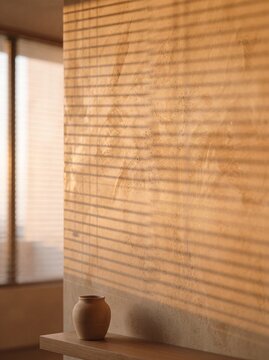 Warm golden hour sunlight through blinds casting shadows on a textured wall with a minimalist clay vase