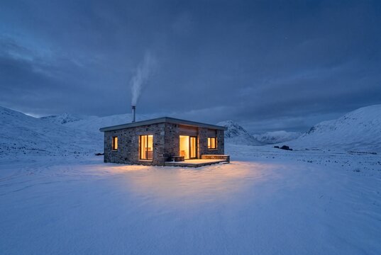 Modern stone cottage in a snowy mountain valley at twilight with warm interior lighting
