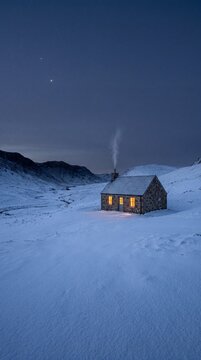 Cozy stone cottage with glowing windows in a snowy mountain valley at twilight