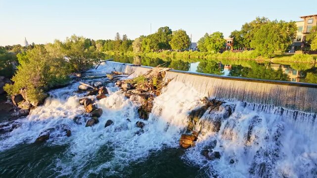 Idaho Falls Temple spire rises above lush trees with Snake River waterfalls cascading in the foreground at sunset