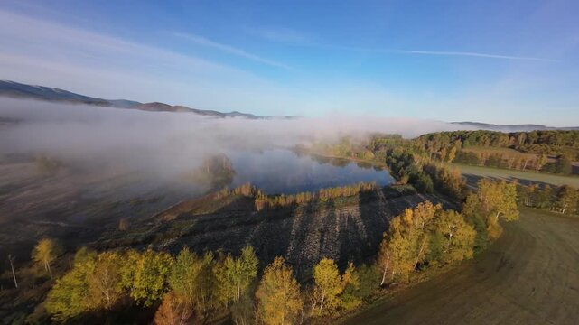 Cinematic aerial drone footage flying through dense morning fog over a peaceful mountain village with small lakes and lush forest. Low clouds cover the valley while the drone rises above the mist
