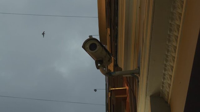 Security camera mounted on building facade under overcast sky with bird flying nearby and power lines