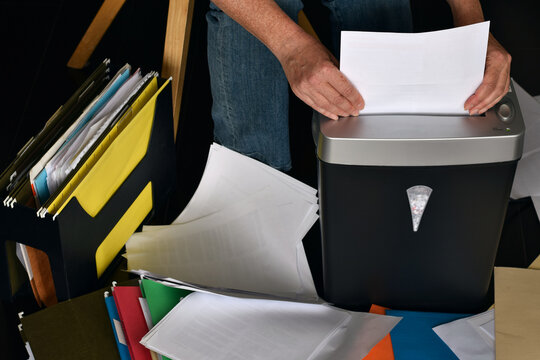 High angle shot showing person feeding white paper into black shredder surrounded by stack of loose document and colorful file folder