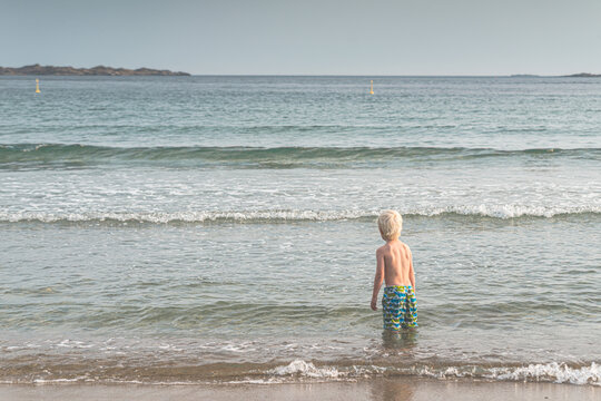 Young blond child wading into shallow sea on sandy beach in serene summer light.
