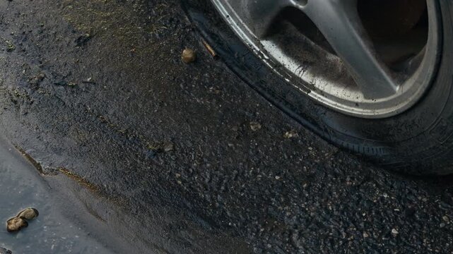 Deflated car tire beside wet pothole on rough asphalt road with damaged wheel rim