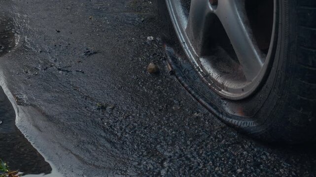 Car wheel rolling through muddy puddle beside rough wet roadway with dark standing water and debris