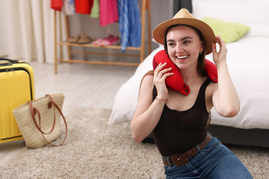 Packing suitcase for trip. Smiling woman in hat with travel pillow on floor at home, space for text