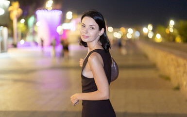 Fototapeta premium At night, smiling girl walks around old city district Barceloneta, in vicinage of Passeig Nou. People are walking by. Person in city, city bustle. Carefree, lively nightlife, promenade. .