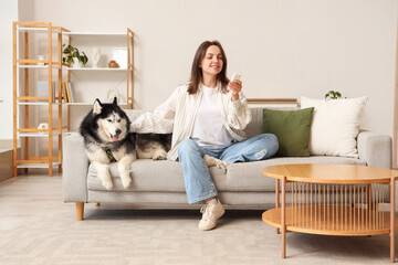 Young woman sitting on sofa with husky dog using remote control © Pixel-Shot