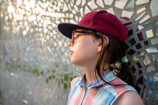 Profile Portrait of a Young Girl Wearing Eyeglassses and a Maroon Snapback Cap Leaning Against a Sparkling Mirror Mosaic Wall Background during Summer