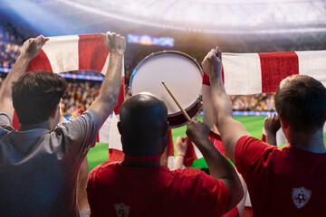 Obraz premium Soccer fans play a drum in stadium tribunes while raising red team scarves, creating passionate energy to support players on the pitch.