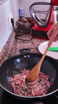 Unrecognizable person stirring sizzling diced pancetta with a wooden spoon in a hot frying pan and covering it with a glass lid, preparing a traditional Mediterranean meal at home