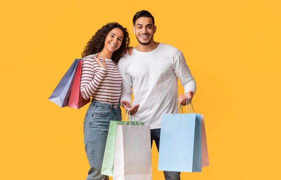 Cheerful Young Middle Eastern Couple Posing With Shopping Bags In Hands Over Yellow Background, Happy Millennial Arab Man And Woman Carrying Purchases, Enjoying Seasonal Sales, Copy Space