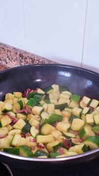 Steaming hot diced zucchini and Italian pancetta saut&eacute;ing in a hot wok pan on an electric stove while a man prepares a traditional Mediterranean pasta sauce in a home kitchen