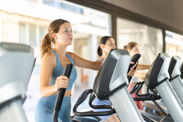 Fototapeta premium Young athletic woman actively training in the gym on the elliptical machine on the platform of other visitors