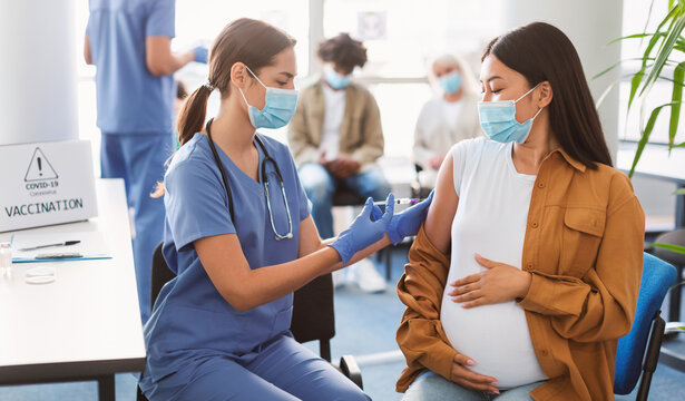 Pregnant asian lady sitting at doctor's office getting modern anti flu shot, hugging belly. Nurse in medical face mask and gloves holding syringe and giving patient vaccine injection in arm